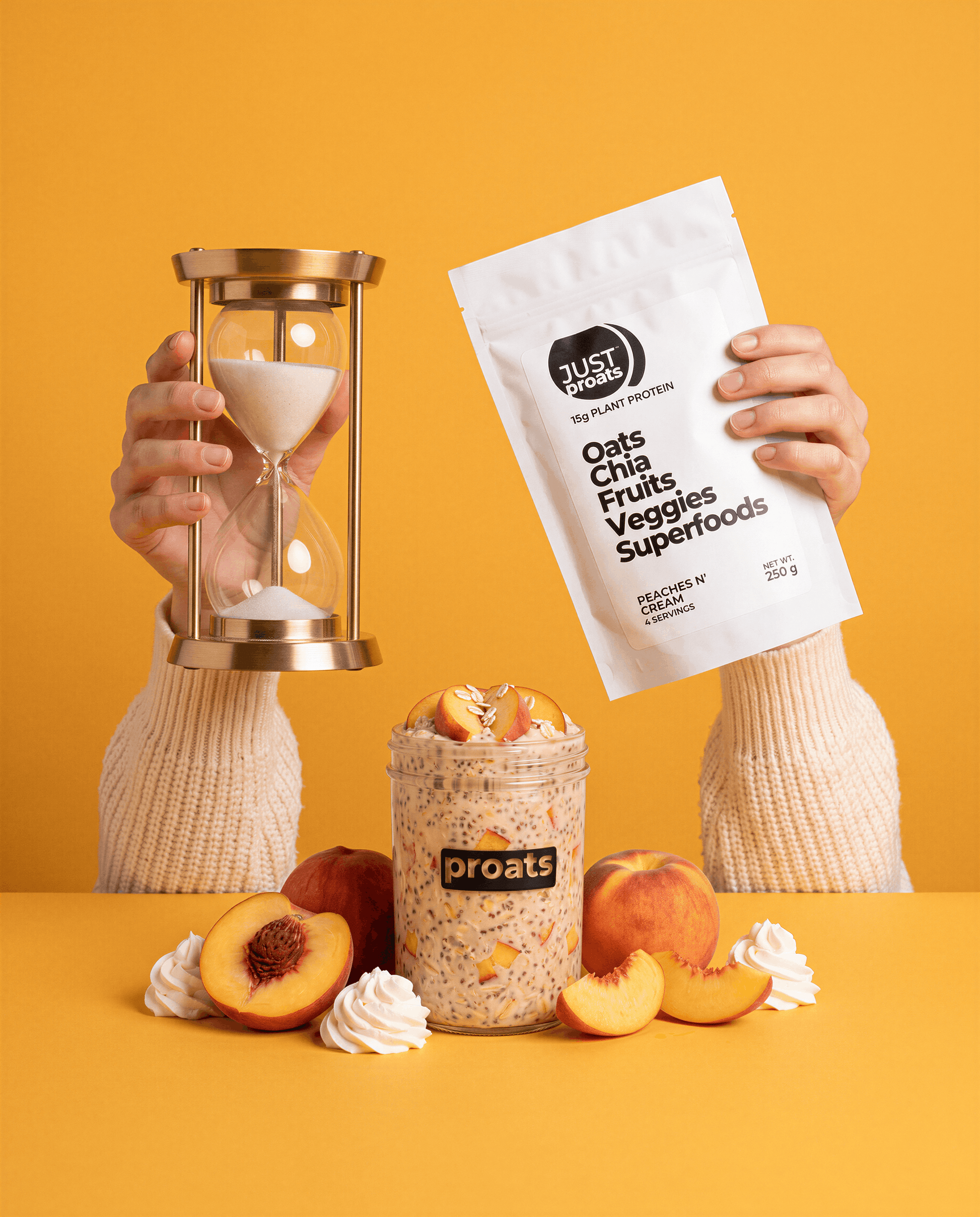 Two hands wearing cream knit sleeves hold a white pouch of Just Proats PEaches n' Cream flavour next to an hourglass. In front sits a jar of Proats overnight oats surrounded by fresh peaches and whipped cream on a solid yellow background.