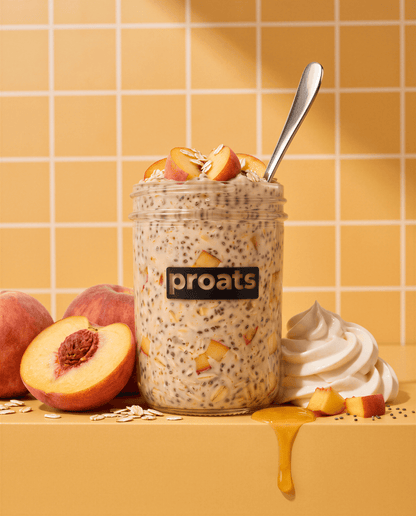A jar of Proats overnight oats topped with peach slices, oats, and a spoon, set on a yellow surface with peaches, whipped cream, and honey against a yellow tiled background.