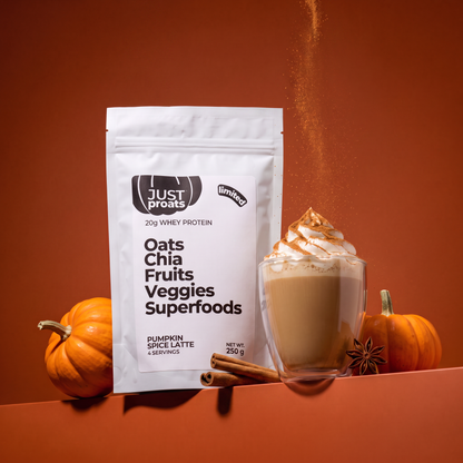 A white stand-up pouch of Just Proats Oats Chia Fruits Veggies Superfoods protein powder, styled next to a glass of pumpkin spice latte with whipped cream and caramel drizzle, with small pumpkins and cinnamon sticks on an orange-brown background.