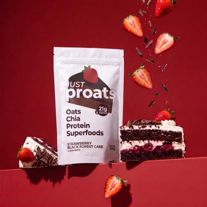 A stand-up pouch of Just Proats Oats Chia Protein Superfoods in Strawberry Black Forest Cake flavor, styled on a dark red background with floating strawberries, chocolate shavings, and two slices of chocolate cake.