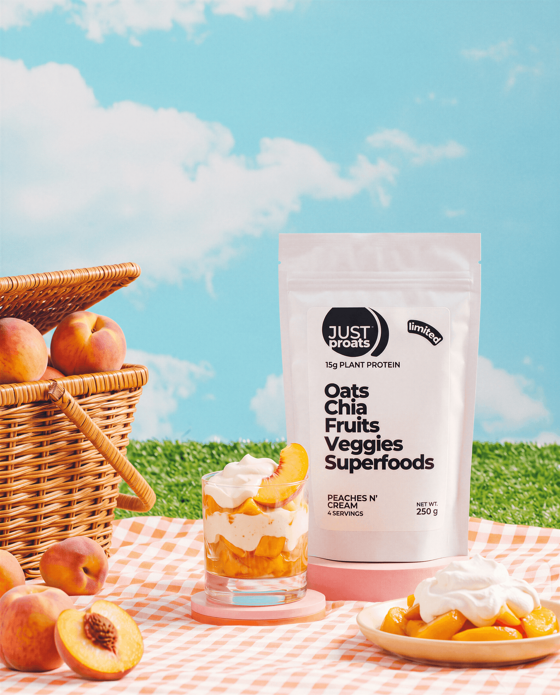 A stand-up pouch of Just Proats on a picnic blanket outdoors. A glass of layered peach and cream drink, a plate of peach slices, and a wicker basket of peaches are arranged around it against a blue sky with clouds background.