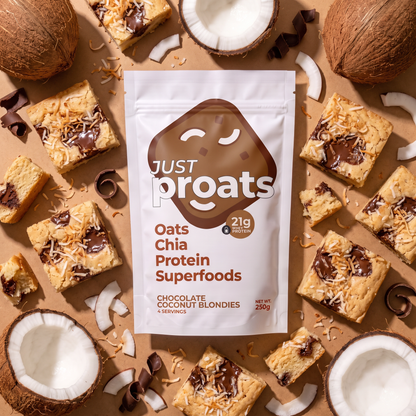 A stand-up pouch of Just Proats chocolate coconut blondies is centered on a tan background, surrounded by sliced blondies topped with chocolate and coconut flakes, plus whole coconuts and coconut pieces.